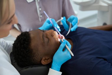 Diş tedavisi sırasında dişçi koltuğunda, profesyonel bakımda yatan Afrikalı Amerikalı bir adama kadın odontolog asistanı.
