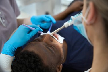 Geçmişe bakış: Afro-Amerikan müşteri dişlerini tedavi eden stomatoloji kliniğinin yüksek nitelikli kişiliği, dişçi anestezi yaparken elinde tükürük püskürtücü tutan hemşire