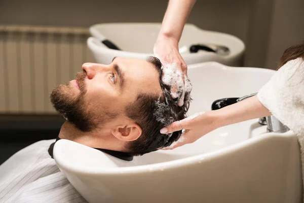 Handsome male enjoys hair washing procedure in barbershop, hairdresser uses special shampoo