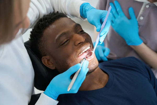アフリカ系アメリカ人の男性クライアントが歯の椅子に横たわり 女性の物理学者が口腔の健康をチェックする口腔鏡と病気のプローブ — ストック写真