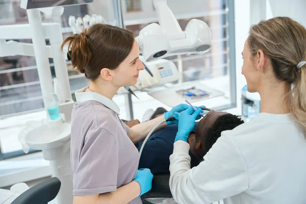 笑顔の看護師は唾液の注射器を保持しながら 女性の専門歯科医は根管療法を維持したり アフリカ系アメリカ人のクライアントに歯を充填したり プロの医療 — ストック写真
