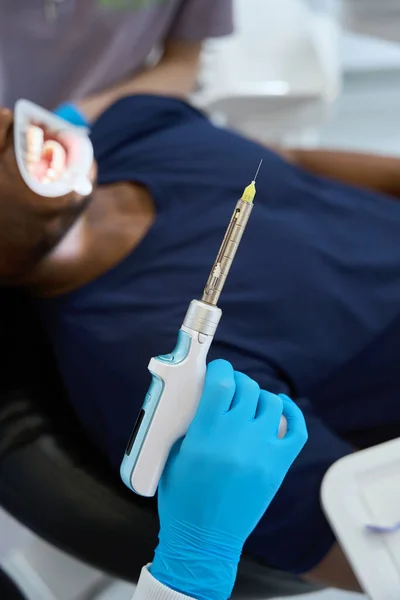 女性の歯科医は 口科医 歯の抽出と歯の椅子に座っている男性のクライアントに痛みのない麻酔を行うために 電気局所麻酔インジェクターを保持しています — ストック写真