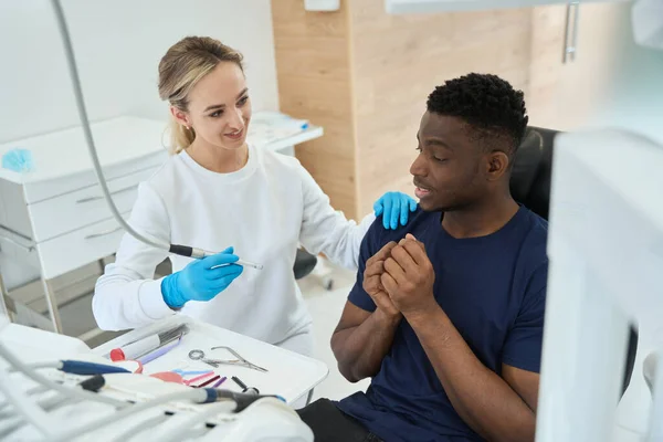 Woman dentist taking dental drill and assuring African American patient that it will not hurt, young man visiting dentist office at first time