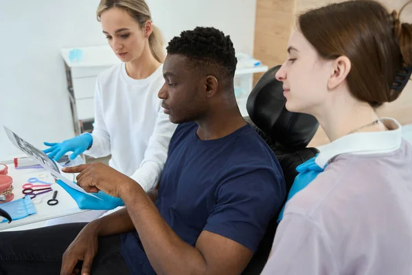 アフリカ系アメリカ人患者がX線画像を見ている彼の歯について質問し 女性の症候学者は問題と治療方法を説明します — ストック写真