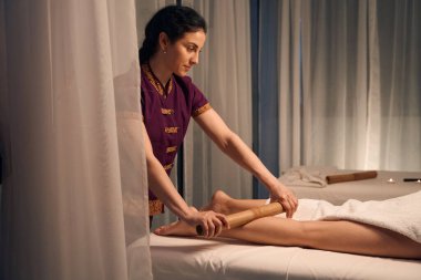 Sakin odaklı masöz kadın hastanın bacağının üzerine bambu çubuğu yuvarlıyor.