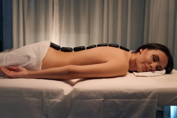 Side view of woman lying prone with smooth stones on her naked back during massotherapy session