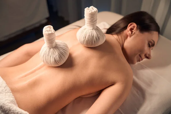 Top view of beauty clinic patient lying on massage bed with herbal bags on her naked back