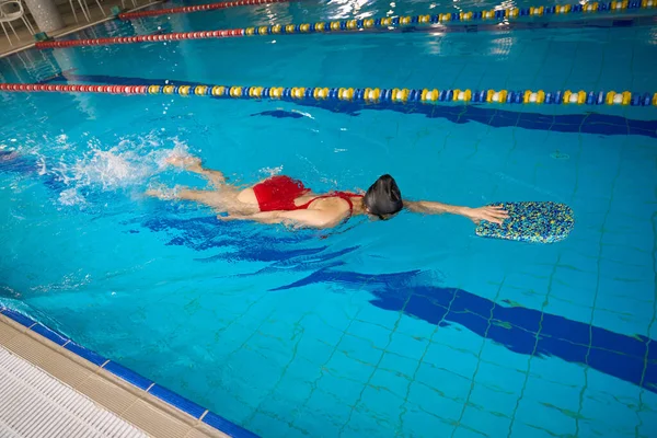Sportswoman in swimsuit and swim cap holding onto kickboard with hand while floating on her stomach in water