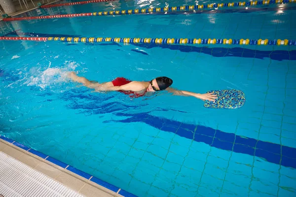 Swimmer in swim cap and goggles holding with hand onto kickboard while swimming front crawl