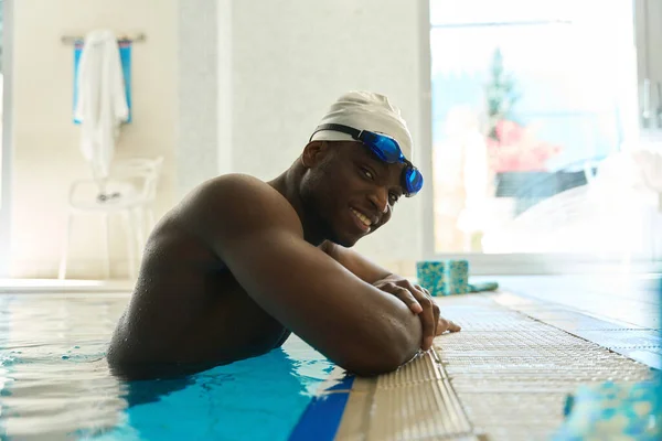 Joyous sportsman in swimming cap and cyan goggles on his head sitting in pool