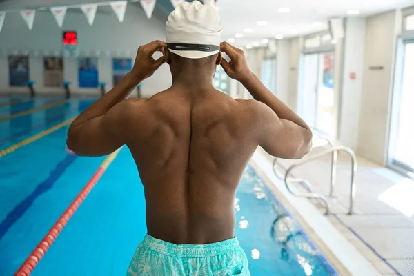 Back view of African American athlete adjusting swim goggles while standing poolside