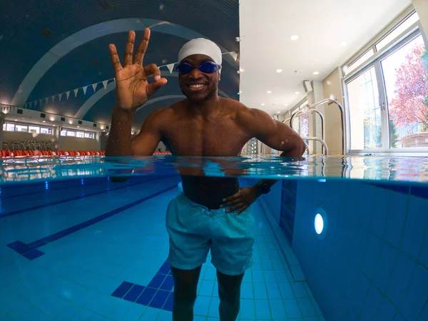 Joyful athletic guy in swim cap and goggles making OK hand gesture while standing in water