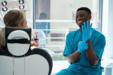 Koruyucu eldivenlerle gülümseyen Amerikalı doktor, klinikte oturan ve dinleyen kadın hasta.