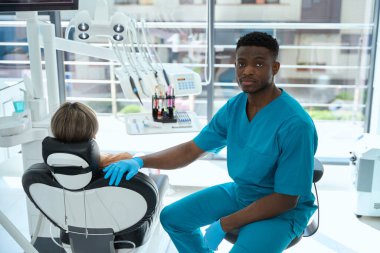 Koruyucu eldivenli ve mavi takım elbiseli bir erkek hastanedeki hastanın yanındaki sandalyede oturuyor ve kameraya bakıyor.