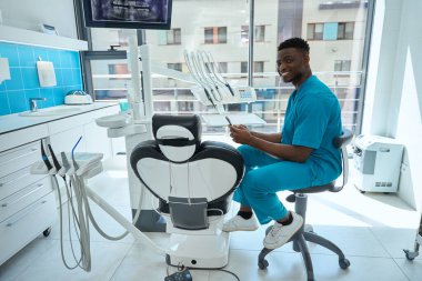 Mavi bornozlu genç doktor, ekipmanın yanındaki sandalyede oturuyor ve elinde telefonla kameraya bakıyor.