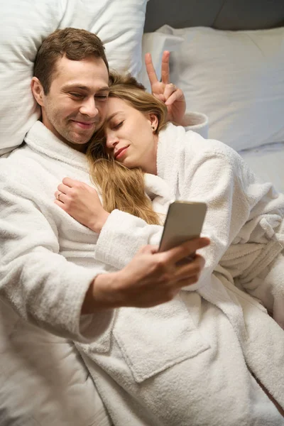 Zabawny Mężczyzna Robi Sobie Selfie Smartfonie Leżącym Szlafroku Łóżku Atrakcyjną — Zdjęcie stockowe