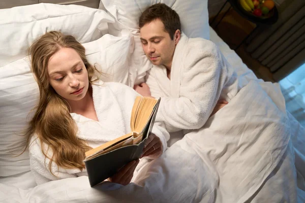 Focused woman reading interesting book while her husband in bathrobe sleeping sweetly lying nearby, misunderstanding between spouses