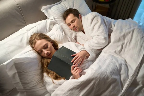 Wife falling asleep reading book lying nearby sleeping husband, couple resting after hard working day in comfortable bed with orthopedic pillows and mattress