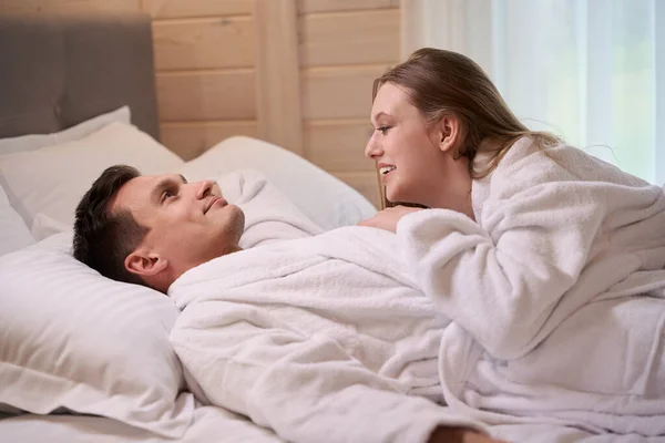 Smiling woman lying on happy man wearing white dressing gowns while staying in bed