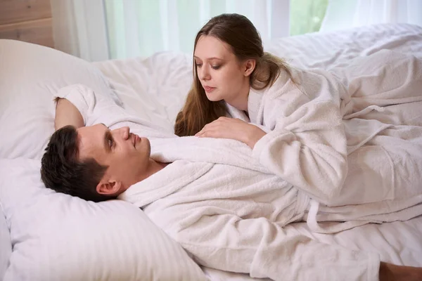 Happy lady lying on smiling man wearing white dressing gowns while staying in bed