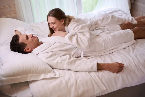 Smiling lady lying on happy man wearing white dressing gowns while staying in bed