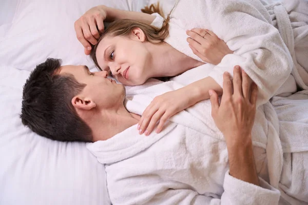 Smiling man and woman in dressing gowns looking at each other while laying in bed