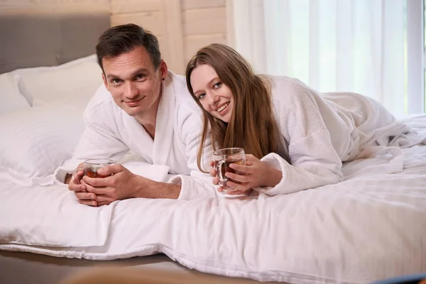 Happy pair in dressing gowns laying in bed and holding teacups in arms