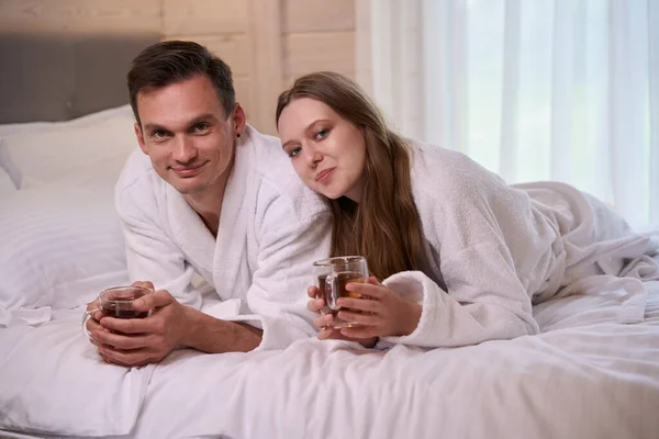 Smiling couple wearing dressing gowns and laying in bed with teacups in arms