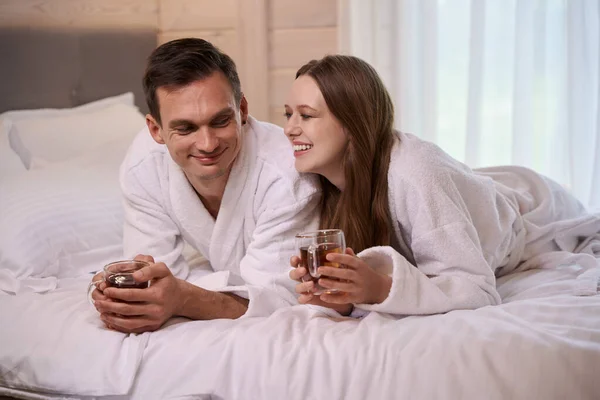 Smiling couple wearing dressing gowns and laying in bed with teacups in arms