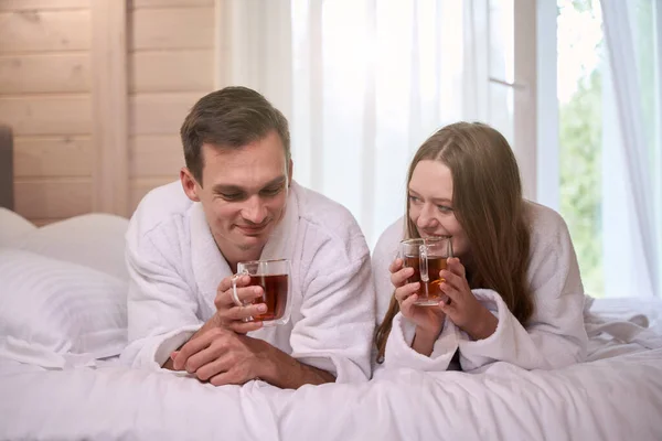 Happy pair wearing dressing gowns and holding teacups while laying in bed