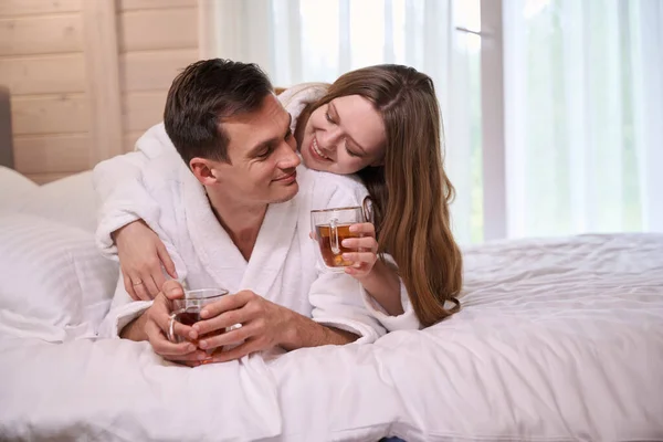 Smilimg pair in dressing gowns lying in bed while holding teacups