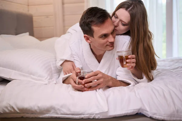 Happy pair in dressing gowns lying in bed while holding teacups