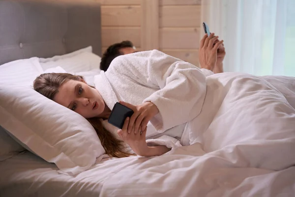 ベッドに横たわってスマートフォンを見ているバスローブで身に着けている驚くべき女性 — ストック写真