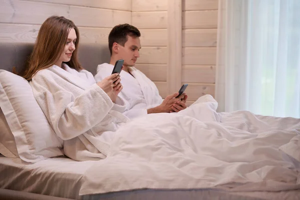 Casal Jovem Vestindo Roupões Banho Deitado Lençóis Brancos Com Telefones — Fotografia de Stock