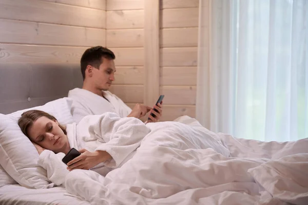 Smiling Couple Wearing Bathrobes Laying White Sheets Phones Hands — Stock Photo, Image
