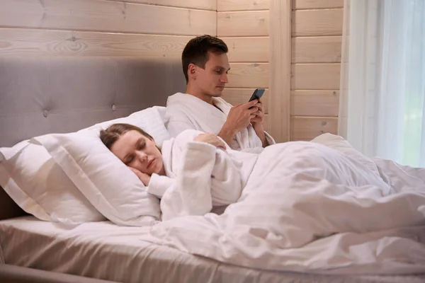 Happy man laying with sleeping lady in bed and looking at smartphone screen