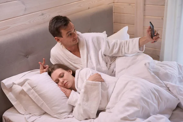 Funny man in dressing gown laying in bed and taking selfie with sleeping lady
