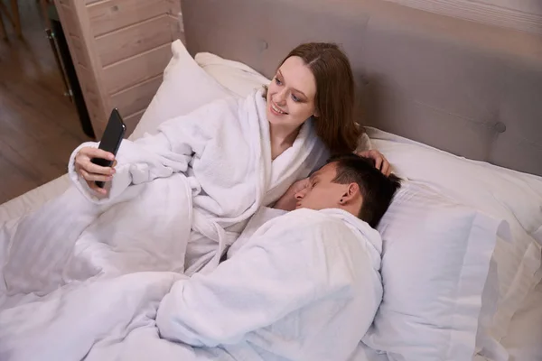 Funny woman in dressing gown laying in bed and taking selfie with sleeping man