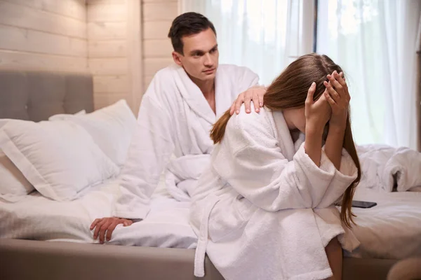 Man comforting upset woman while sitting on bed in white dressing gowns