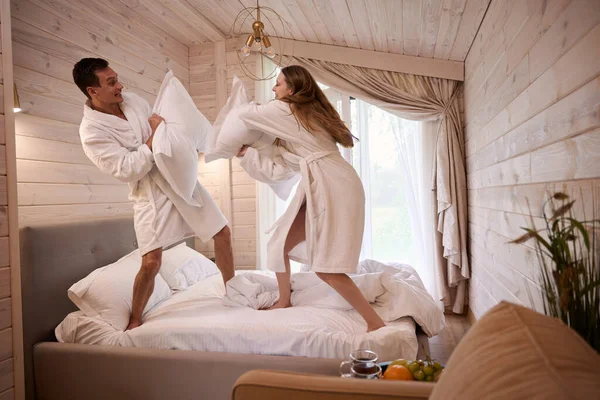 Happy couple wearing dressing gown and having fun pillow fight on bed