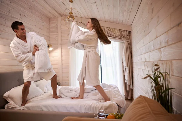 Smiling couple in dressing gown having fun while fighting with pillows in bedroom