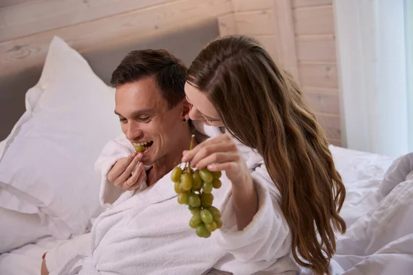 Woman wearing dressing gown and feeding happy man with grapes in bed