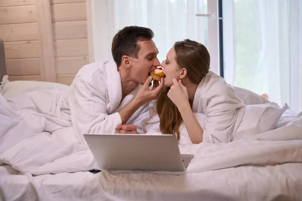 Birlikte Pasta Yerken Bornozlu Bir Çift Dizüstü Bilgisayarla Yatakta Yatıyor — Stok fotoğraf