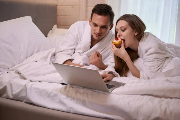 Couple Bathrobes Laying Bed Laptop While Woman Eating Cake — Stock Photo, Image