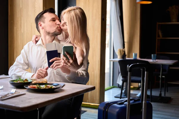 Beautiful couple tenderly kissing holding passports and tickets and making selfie on smartphone, suitcase standing nearby, people waiting transfer on cafe near airport