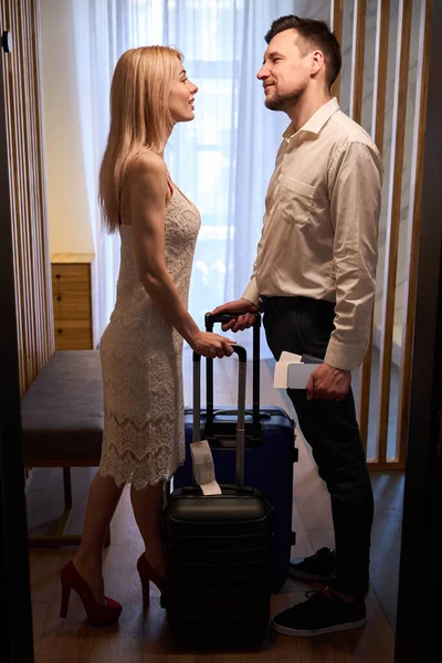 Happy woman in white dress going to kiss her husband, couple standing in hotel room with suitcases, hotel check-in, honeymoon celebration abroad
