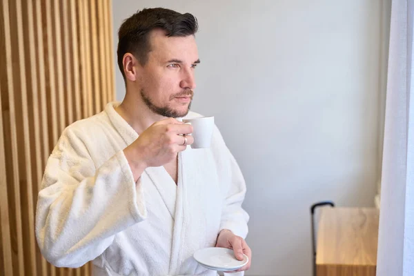 身穿保暖的白色浴衣 头戴胡子的男人 早上喝新鲜煮好的咖啡 看着窗户 工作前精力充沛 — 图库照片