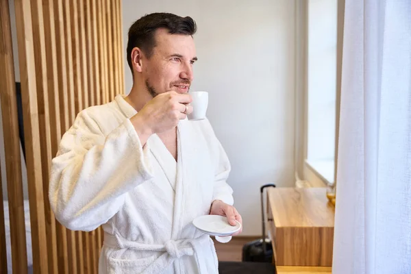 微笑的男人穿着柔软的浴衣 喜欢早上喝咖啡 看窗户 出差和住在宾馆里 — 图库照片