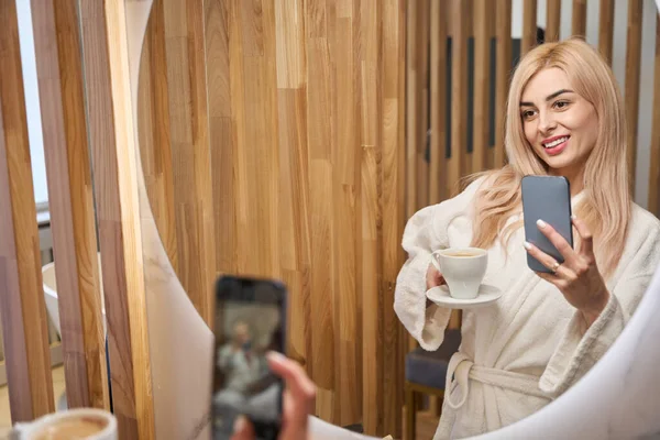 迷人的金发女人穿着柔软的浴衣 拿着咖啡 在酒店房间的镜子里的智能手机上自拍 在社交网站上发布 — 图库照片