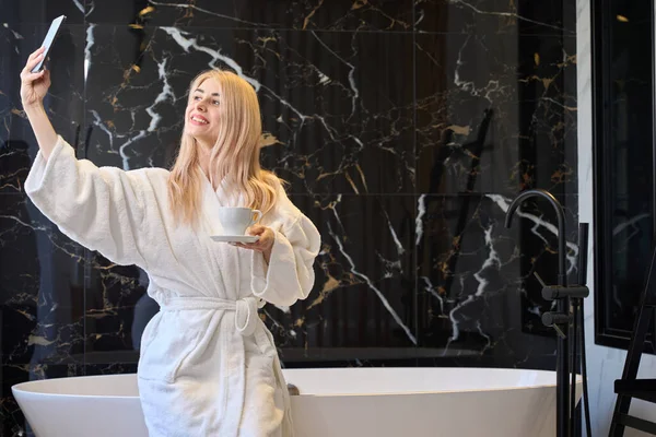 一个积极快乐的女人 带着豪华宾馆的公寓在浴室里做浴衣和喝咖啡 自夸一番 — 图库照片